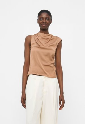 Femeie purtând un top bej din satin fără mâneci și pantaloni albi cu talie înaltă, stând în fața unui fundal simplu și deschis la culoare.