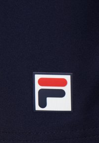Le tissu bleu marine présente un logo rectangulaire blanc avec des accents rouges et bleus, affichant des lettres stylisées "F" et "L", détaillées avec des rayures verticales.