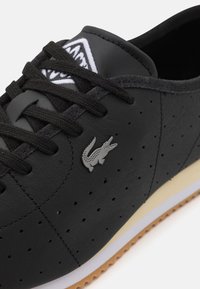 Fekete lyukacsos bőr sneaker fekete fűzőkkel, ezüst krokodil logóval, fehér középtalppal és gumi talppal.