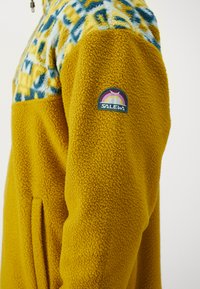Gul fleecejakke med et mønstrete blått og gult øvre parti. Har et SALEWA-logo-merke på ermet og side lommer.