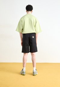 Camisa de manga corta de gran tamaño en verde claro y pantalones cortos negros. El modelo lleva zapatillas grises con detalles en verde. Diseño simple sin patrones visibles.
