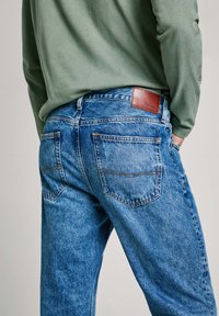 Los jeans de mezclilla azul claro presentan un corte clásico, costuras en contraste y un parche de cuero marrón en la cinturilla con el logotipo de la marca.