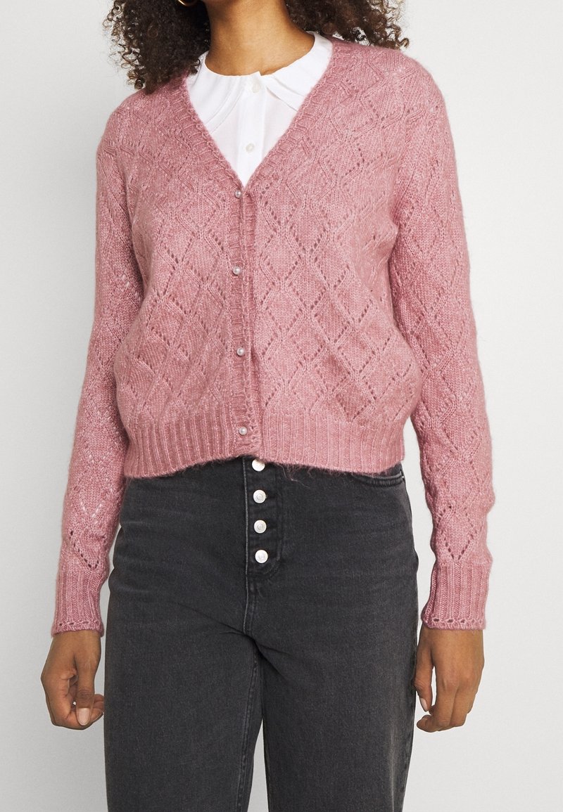 Žena v růžovém úpletovém kardiganu s perlovými knoflíky přes bílou košili s límečkem a vysokým pasem černými džínami s knoflíkovým zapínáním.