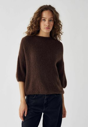 Pull en tricot texturé marron avec un col rond et des manches courtes bouffantes, porté sur un jean foncé devant un fond clair.