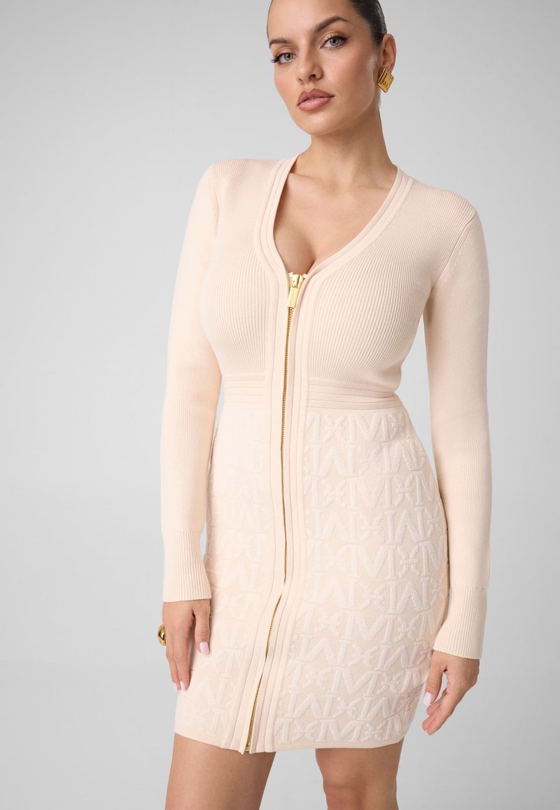 Femme portant une robe beige côtelée zippée avec des lettres en relief, une fermeture éclair dorée et des boucles d'oreilles dorées, debout devant un fond clair uni.