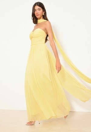 Kvinde i flydende, skulderfri gul kjole med lang tørreldetalje, der går mod en ensfarvet hvid baggrund.