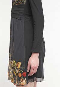 Czarna sukienka z długimi rękawami, z marszczonym biustem, transparentną nakładką oraz kwiatowym wzorem w żółtym, pomarańczowym i zielonym na dolnym brzegu. Gładka faktura.