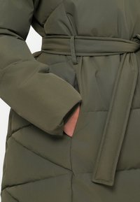 Olivgrüner gesteppter Steppmantel mit passendem Stoffgürtel und einer Hand, die in eine Seitentasche gesteckt ist.