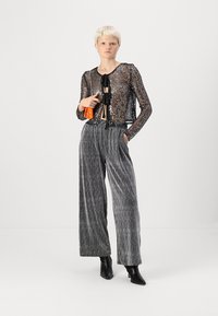 Črna prosojna srajca z dolgimi rokavi in bleščicami, v pari z visokim pasom in širokimi sivimi črtastimi hlačami. Model drži oranžno torbico in nosi črne čevlje.