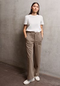 Chemise blanche à manches courtes associée à un pantalon large marron neutre avec des poches avant et une fermeture à bouton. Des baskets blanches complètent le look.