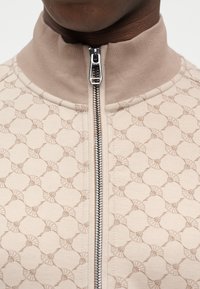 Veste beige zippée avec un motif texturé. Dotée d'un col montant et d'une fermeture éclair argentée. Le matériau a un léger éclat.