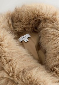 Faux fur krave i lyse beige, med en blød, fluffy tekstur. Indvendigt er der et glat stof med en label, der læser "NEXT," hvilket angiver mærke og pleje.