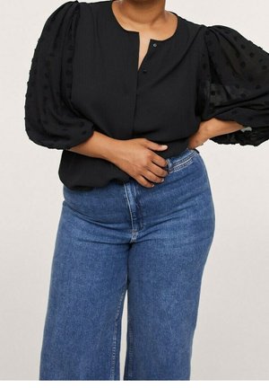 Personne portant un chemisier noir avec des manches bouffantes texturées et un jean bleu taille haute à jambes larges, debout avec une main sur la taille.