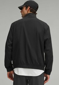 Chaqueta ligera negra con cuello alto, puños elásticos y banda en la cintura. Presenta una textura suave y un diseño minimalista. Combinada con una camisa blanca.