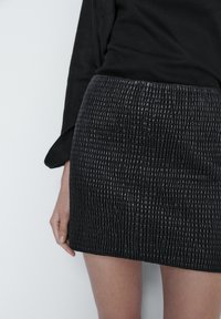 Zwart textured mini-jurk met een reliëfpatroon, gemaakt van een glanzend materiaal, gecombineerd met een strakke zwarte top, waarbij een deel van de hand zichtbaar is.