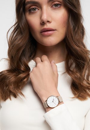 Ronde horloge met een witte wijzerplaat, gouden accenten en een lichtgrijze leren band, gedragen om een pols. De drager heeft een witte top aan en golvend haar.