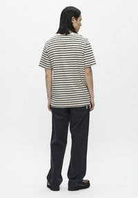 Tricou bej în dungi, cu mâneci scurte, asociat cu pantaloni negri închis. Tricoul are dungi orizontale negre și o etichetă mică cu logo.