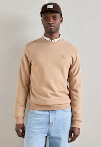 Felpa beige con tessuto testurizzato, colletto rotondo e polsini a costine. Abbinata a jeans in denim azzurro chiaro. Cappello marrone con dettagli del logo.