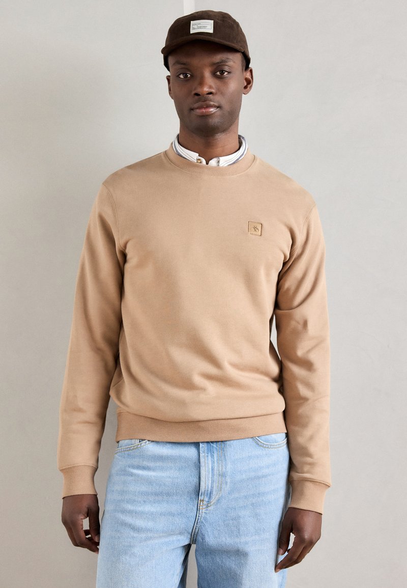 Felpa beige con tessuto testurizzato, colletto rotondo e polsini a costine. Abbinata a jeans in denim azzurro chiaro. Cappello marrone con dettagli del logo.