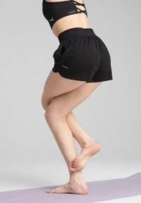 Mustad mustad lühikesed püksid lõdva lõikega, elastse vöökohaga, brändi etikett. Mudel poseerib heledal lillal vaibal, paljajalu.