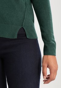 Pull vert en maille avec poignets et ourlet côtelés, doté de fentes latérales en bas, associé à un jean bleu foncé.