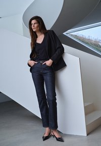Woman in black blazer, dark jeans, and black heels leaning against white modern staircase railing indoors.