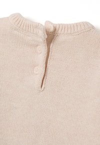 Close-up van een licht beige gebreide trui met een geribbelde kraag en drie kleine bijpassende knopen die gedeeltelijk zijn vastgemaakt.
