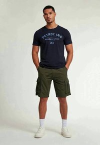 Marineblauwe katoenen t-shirt met witte en blauwe tekst, gecombineerd met olijfgroene cargo shorts met zijzakken en witte sneakers.