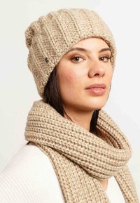 Beanie beige in maglia con una trama a coste, caratterizzato da un piccolo accento metallico. Abbinato a una sciarpa chunky in maglia beige coordinata.