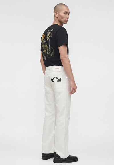 Άνδρας που φοράει μαύρο μπλουζάκι με λουλουδάτο γραμμικό σχέδιο στην πλάτη, άσπρο τζιν με μαύρο λογότυπο βέλους στην τσέπη και μαύρα παπούτσια, στέκεται πλάγια.