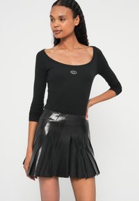 Μαύρο ριμπ μακρυμάνικο τοπ με στρογγυλή λαιμόκοψη και λεπτομέρεια λογότυπου, συνδυασμένο με μαύρη λαμπερή πλισέ μίνι φούστα από ψεύτικο δέρμα.
