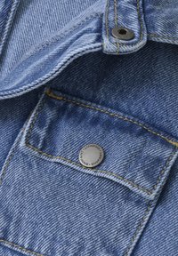 Denimtyg i ljusblått med en bröstficka och synlig gul sömnad. Har en silverfärgad metallknapp med ingraverad text.