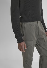 Crni pletivni pulover s teksturom, uparen s sivim cargo hlačama koje imaju više džepova i elastičnu maramu. Prikaz izbliza sa strane.
