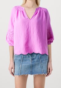 Blusa rosa, leggera, con scollatura a V, tessuto testurizzato e orlo scalloppato. Presenta dettagli ricamati sulle maniche a sbuffo. Abbinata a una gonna di jeans.