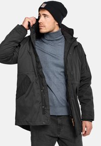 Chaqueta de invierno negra con cremallera en la parte delantera, grandes bolsillos y capucha, llevada sobre un cuello de tortuga gris. Tejido suave y puños ajustables.