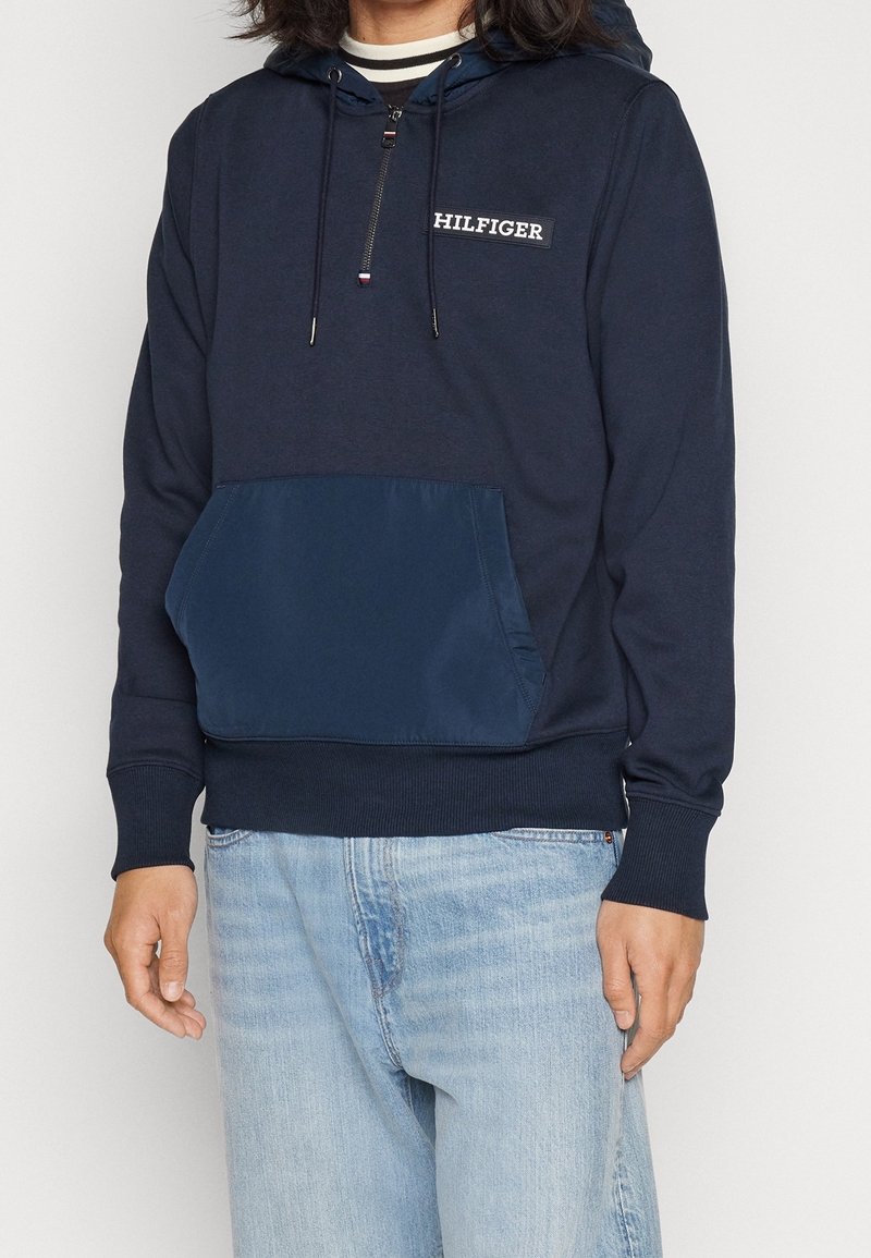 Homme portant un sweat à capuche bleu marine avec une fermeture éclair sur le devant, une grande poche frontale et un logo "HILFIGER", assorti à un jean bleu clair.