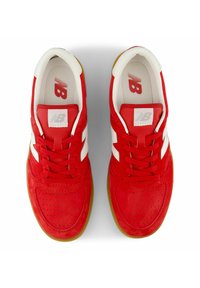 Red suede sneakers feature white accent stripes, perforated detailing, and a gum rubber outsole. Iconic logo is visible on the tongue.