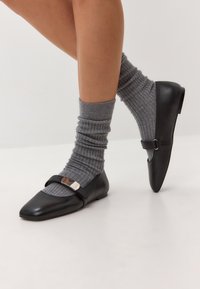 Balerini negri din piele, cu vârful ascuțit, având o curea și detalii cu cataramă metallică, purtați cu șosete gri cu dungi. Textură netedă.