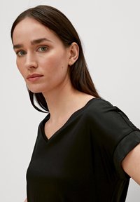 T-shirt nera a V con tessuto liscio, caratterizzata da maniche corte arrotolate e design minimalista, indossata da una donna con capelli lunghi.