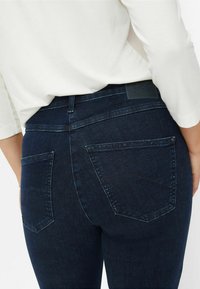 Jean en denim bleu foncé avec une taille mi-haute, deux poches arrière et une étiquette texturée sur la taille. Matériau extensible pour le confort.