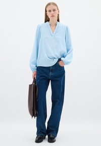 Ljusblå, långärmad blus med v-ringning och texturerad tyg, i kombination med högmidjade mörkblå raka jeans. Brun axelväska i handen.