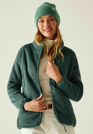 Kobieta w turkusowej dzianinowej czapce, turkusowej polarowej kurtce na zamek, kremowym golfie i jasnych spodniach, poprawiająca zamek kurtki.