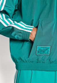 Lätt turkos jacka med vita ränder på ärmarna, elastiska muddar och en ficka med en patch med Liverpool Football Club-logotyp.