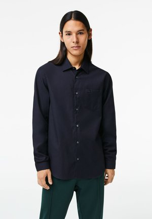 Camisa de botón de manga larga en azul oscuro, con un bolsillo en el frente y una textura de tela suave. Presenta un cuello y un sutil detalle de botones.