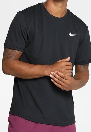 Mężczyzna w czarnej koszulce Nike z krótkim rękawem trzyma lewe nadgarstek prawą ręką, pokazując tatuaże na ramionach.