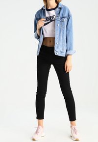 Person som bär en kort vit Nike-tröja, ljusblå denimjacka, svarta skinnybyxor och rosa sneakers stående mot en vit bakgrund.