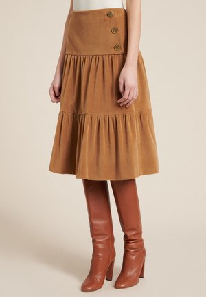 Femme portant une jupe marron à étages longueur genou avec trois boutons décoratifs et des bottes en cuir marron montantes à talons.