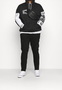 Felpa nera con testo e grafiche bianche, abbinata a pantaloni neri e sneakers bianche. Presenta una borsa a tracolla nera opaca e elegante.