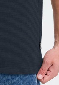 Tamsiai mėlynas, tekstūruotas audinys marškiniai su žyma šone. Rankos sukabinta apačioje šalia džinsų, rodomas marškiniai detalizavimas ir kirpimas.