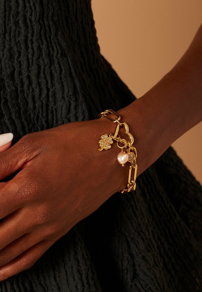 Gouden schakelarmband met een klavertjevier charm met heldere stenen en een witte parel pendant, rustend op een pols tegen een gestructureerde donkere stof.